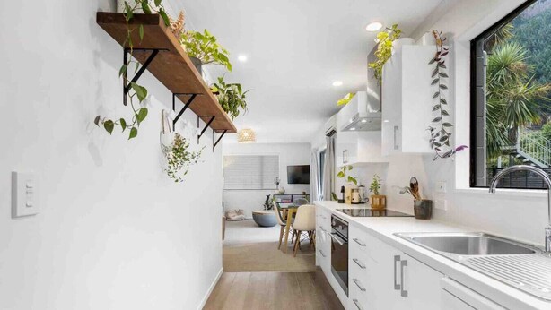 Una cocina moderna con armarios blancos, un estante de madera con plantas, un comedor con mesa y sillas, y una ventana con vistas al verdor.