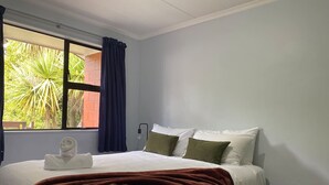 A bedroom with a bed, a window with curtains, a bedside table, and a view of greenery outside. - Robins Retreat-Town Center - Parking - Easy access (Queenstown)