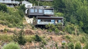 Uma propriedade costeira moderna com vários edifícios, incluindo uma casa de destaque com telhado inclinado e uma construção menor com telhado plano. A propriedade está localizada numa encosta coberta de vegetação densa e com um corpo de água ao primeiro plano.
