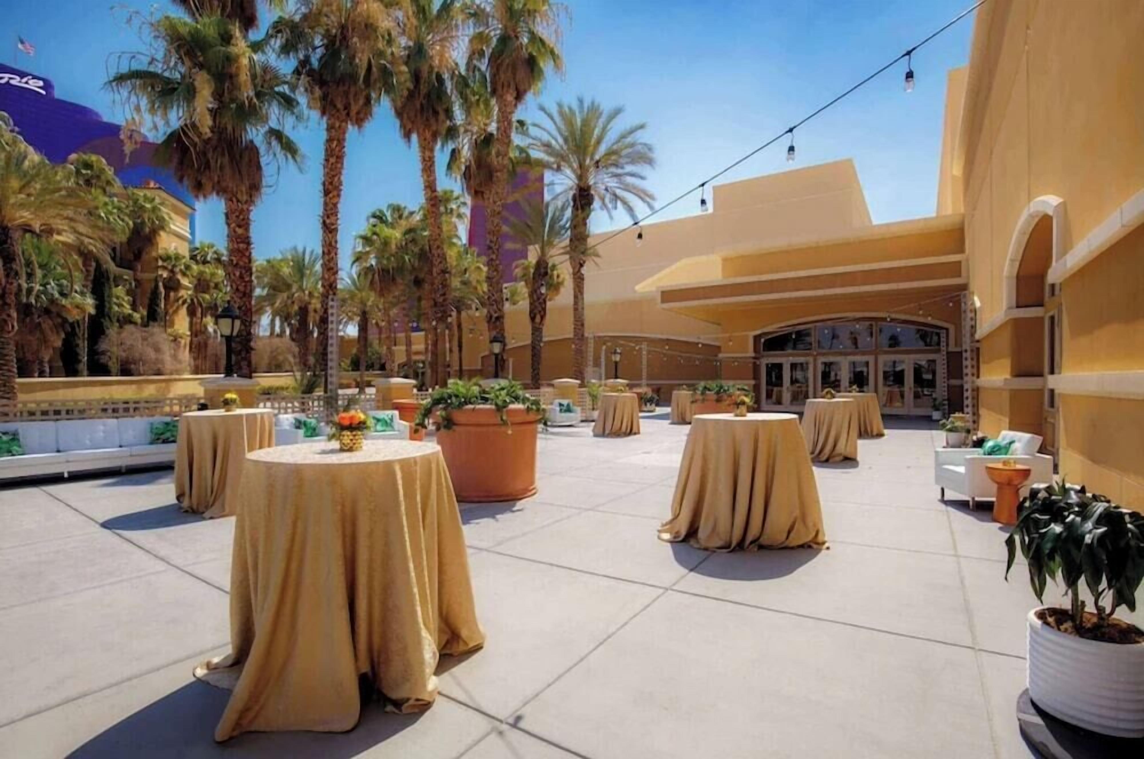 An outdoor patio area with round tables covered in beige tablecloths, surrounded by potted plants and palm trees.