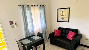 A living room with a black sofa, a dining table with two chairs, a framed picture on the wall, and a window with curtains. - SeaEsta Negril (Negril)