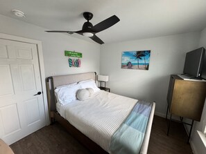 A bedroom with a bed, a ceiling fan, a painting of a beach, a sign on the wall, and a television on a stand.
