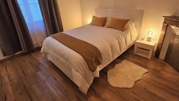A bedroom with a large bed, wooden flooring, a nightstand with a lamp, and a window with curtains.
