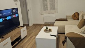 A modern living room with a flat-screen TV, a white coffee table, a sofa, and a window with sheer curtains.