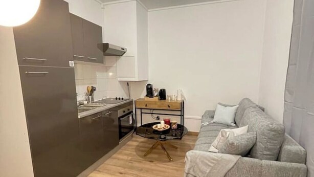 Et lille køkken med moderne hårde hvidevarer, et spisebord med enkelte ting og en grå sofa i et kompakt opholdsområde.