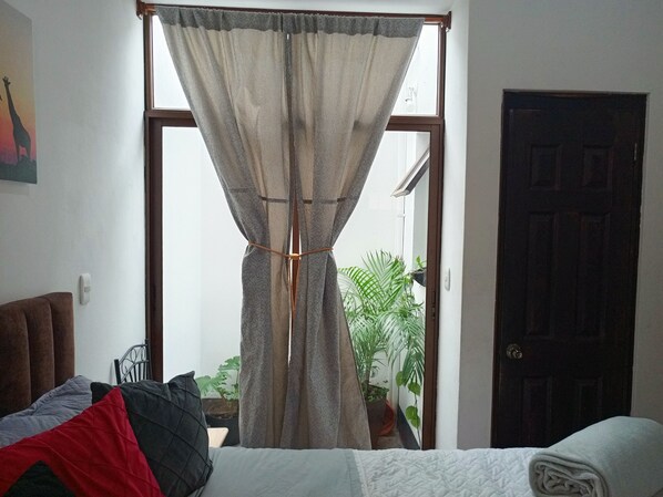 A room with a bed, a dark wooden door, a window with curtains, and a wall-mounted thermostat.