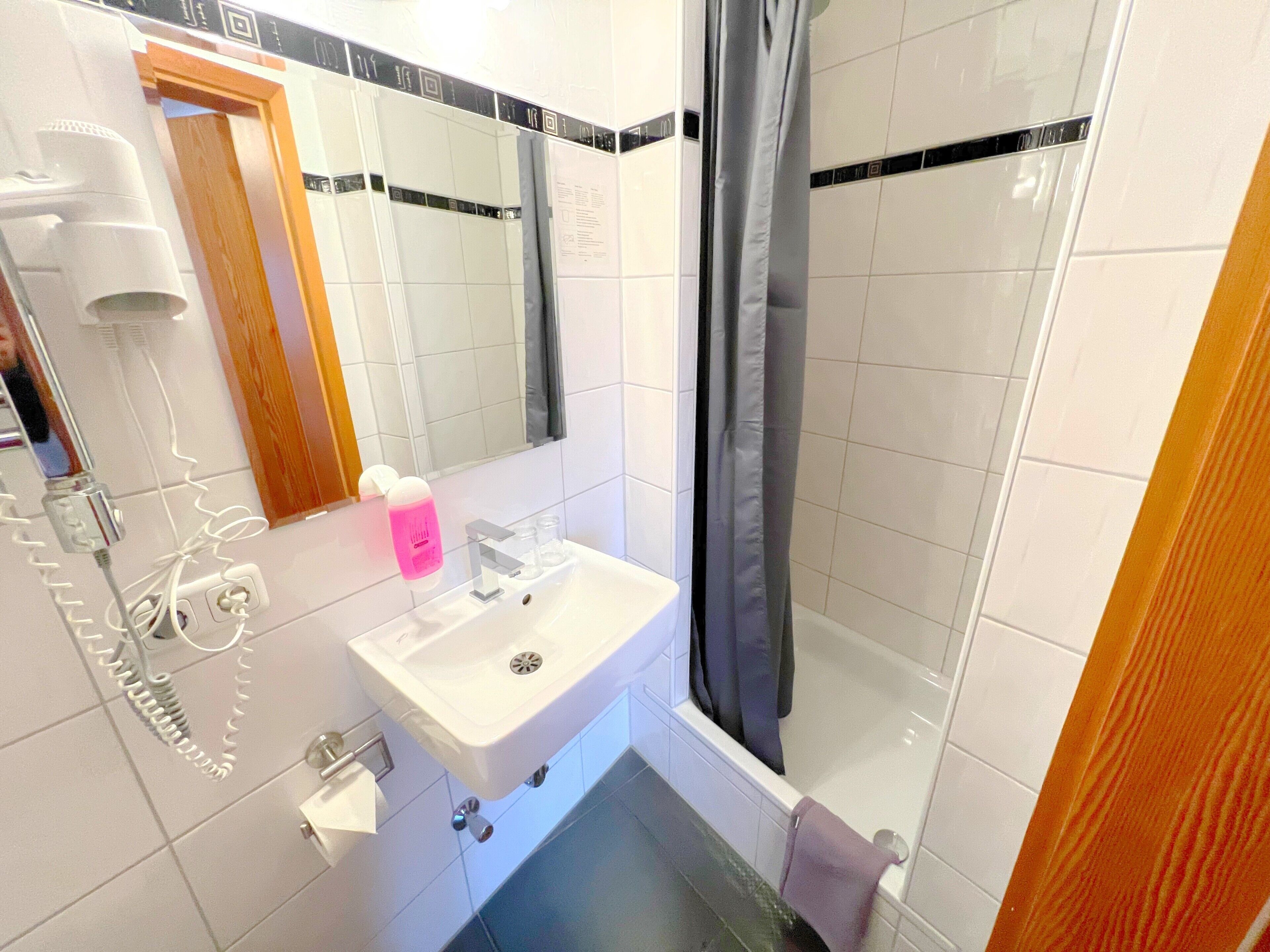 A bathroom with a sink, mirror, shower, and a hairdryer on the wall.