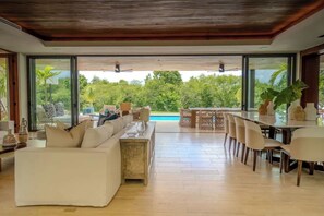 Un vaste espace de vie comprenant un canapé, une table à manger et des chaises, de grandes fenêtres offrant une vue sur la verdure et une piscine.