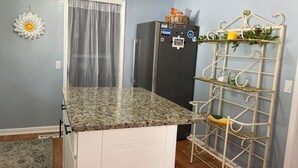 A kitchen with a granite countertop, a refrigerator, a white cabinet, a decorative metal rack, and a window with sheer curtains. - Hillside Hideaway (Weber City)
