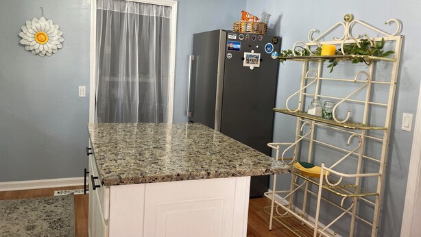 A kitchen with a granite countertop, a refrigerator, a white cabinet, a decorative metal rack, and a window with sheer curtains. - Hillside Hideaway (Weber City)
