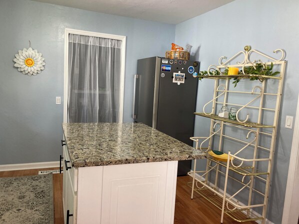 Una cocina con encimera de granito, refrigerador, un armario blanco, una estantería metálica decorativa y una ventana con cortinas translúcidas.