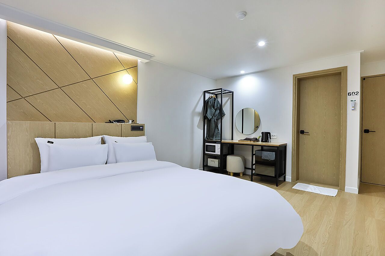 A modern hotel room with a large bed, a wooden headboard, a desk with a mirror, and a wardrobe.