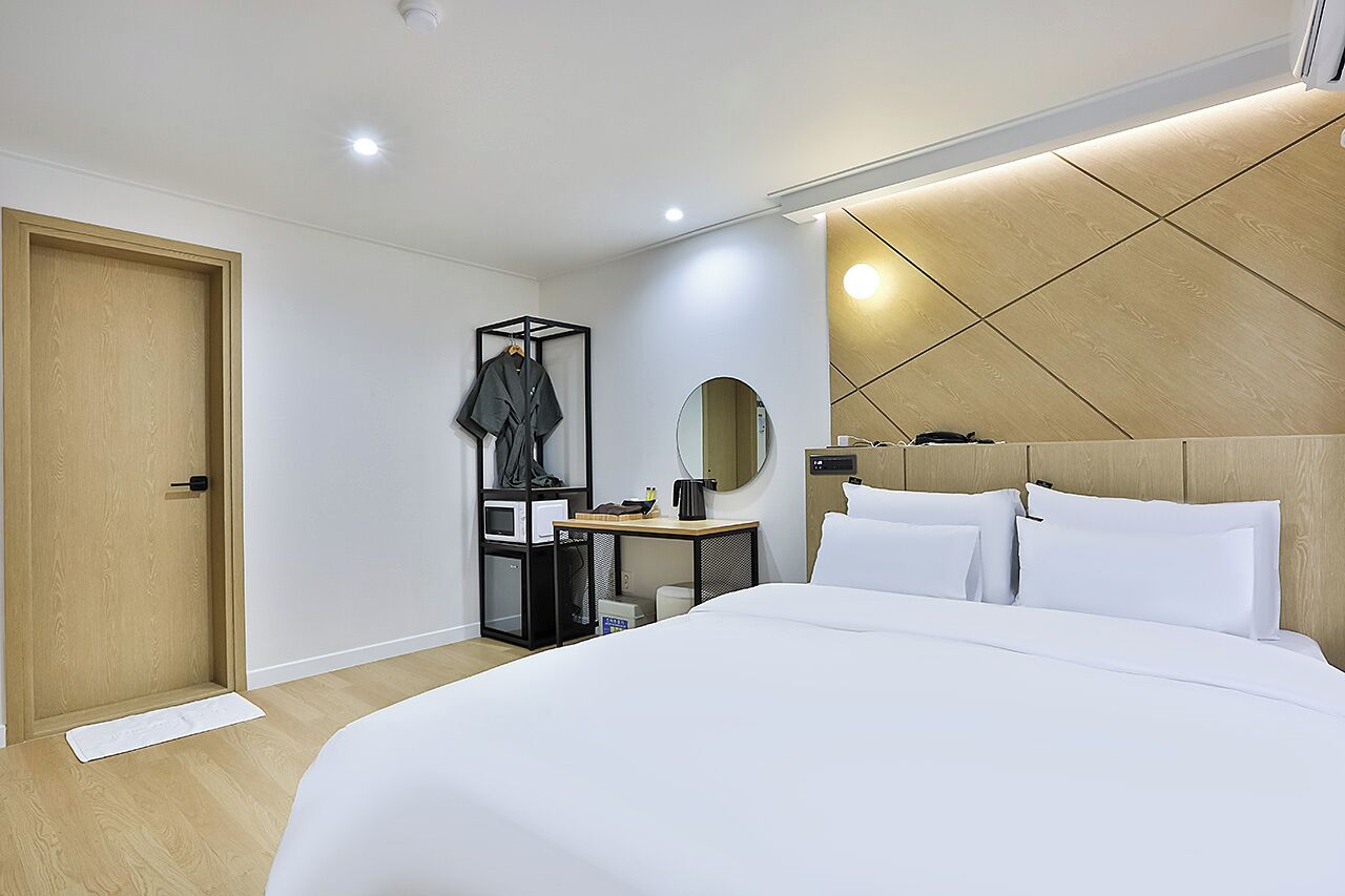 Una habitación de hotel moderna con una cama grande, una puerta de madera, un perchero, una mesita con un espejo y una lámpara fijada a la pared.