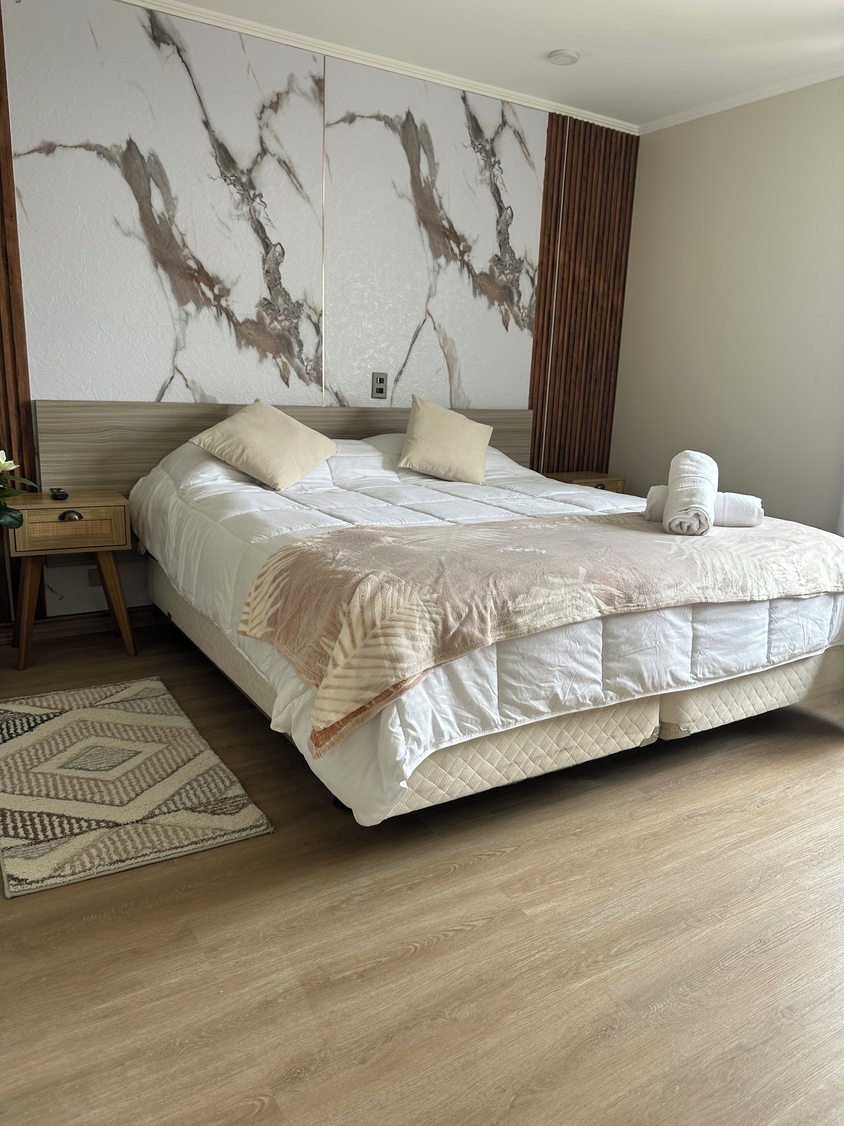 A modern hotel room with a large bed, a wooden headboard, a marble wall, and a wooden floor.
