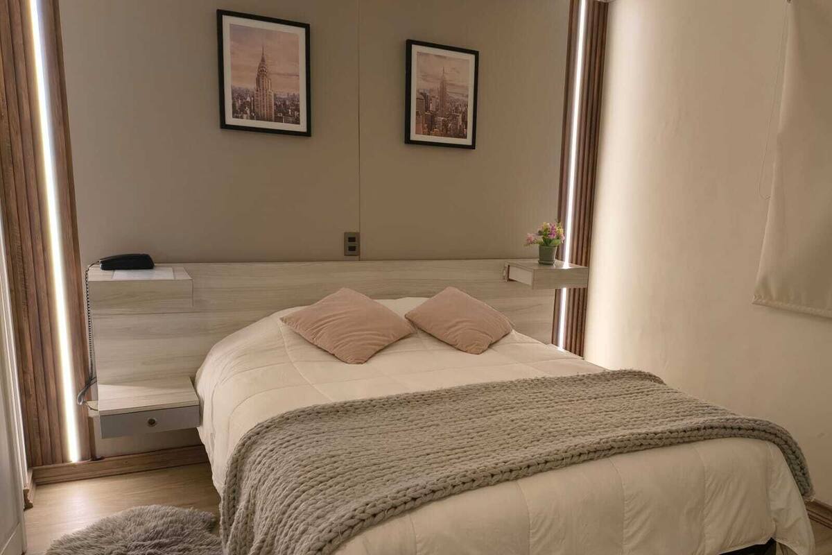 A bedroom with a bed, a bedside table, a lamp, a mirror, and two framed pictures on the wall.
