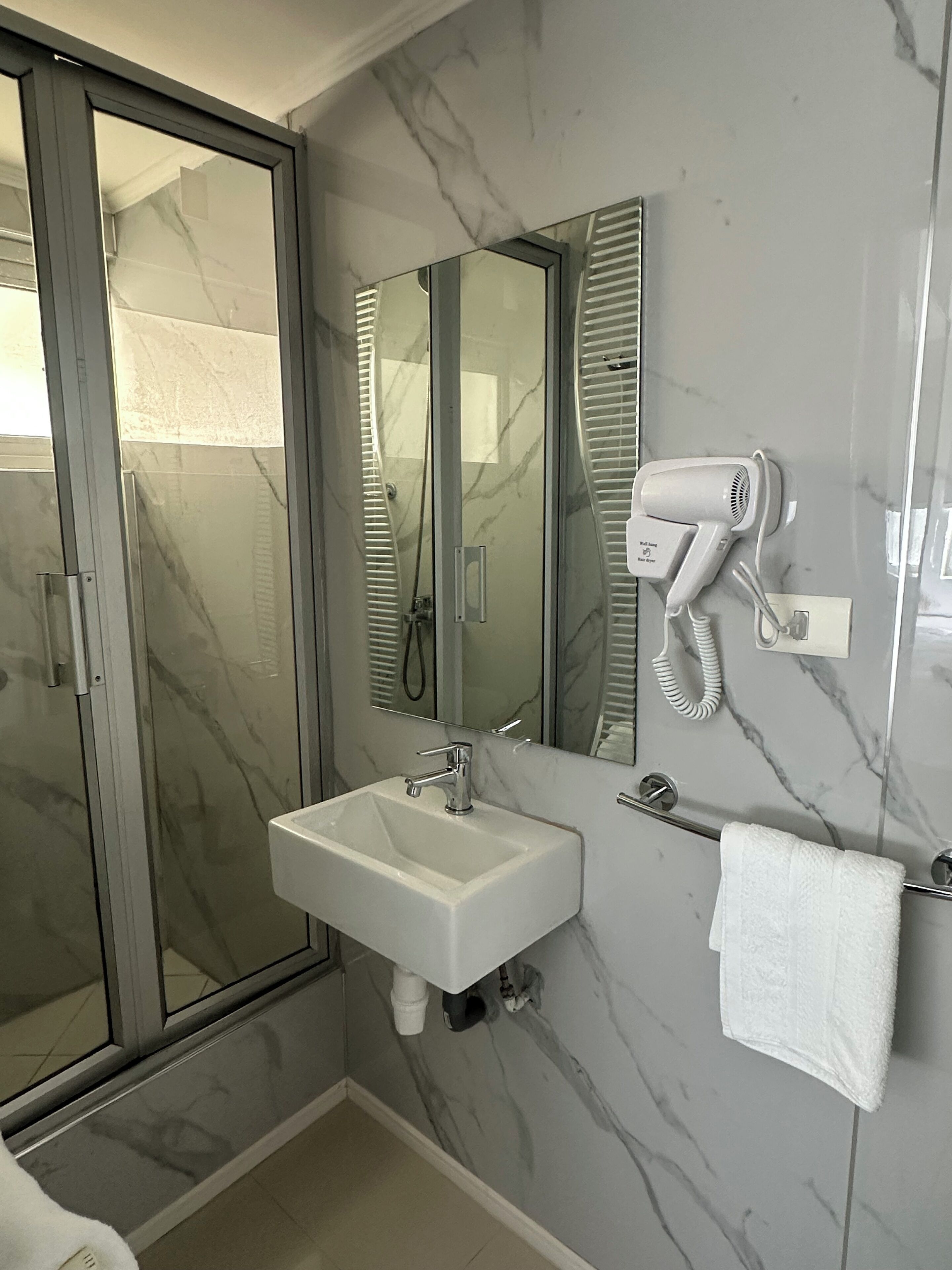 A modern bathroom with a glass shower enclosure, a white sink, a mirror, and a hairdryer mounted on the wall.