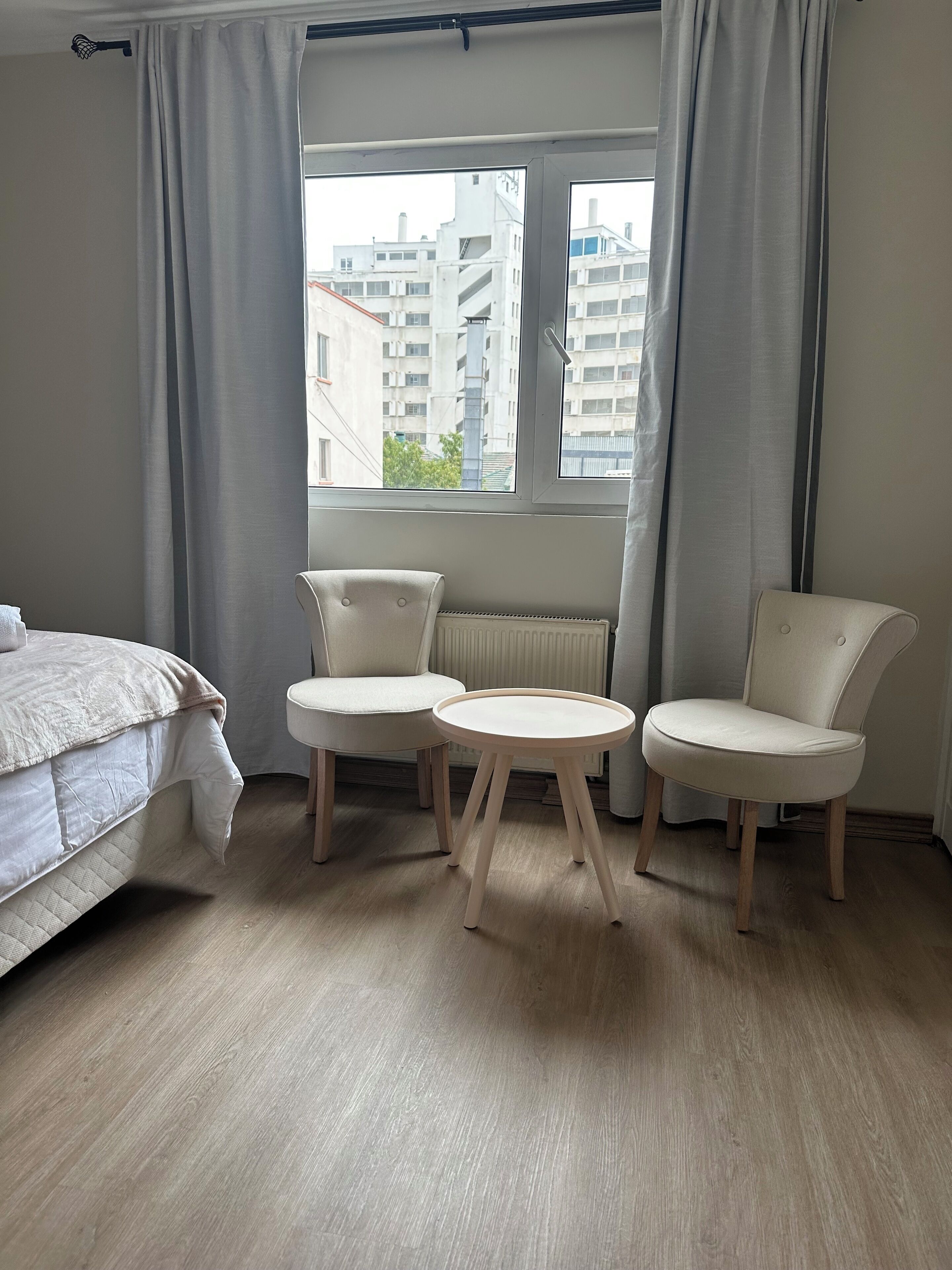 A bedroom with a bed, two chairs, a small table, a window with curtains, and a radiator.