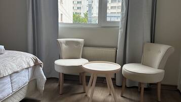 A bedroom with a bed, two chairs, a small table, a window with curtains, and a radiator.