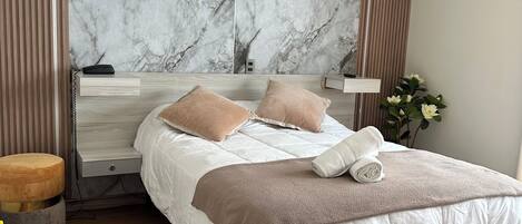 A modern bedroom with a marble-patterned headboard, a wooden bedside table, a round ottoman, a patterned rug, and a neatly made bed with a brown throw blanket.