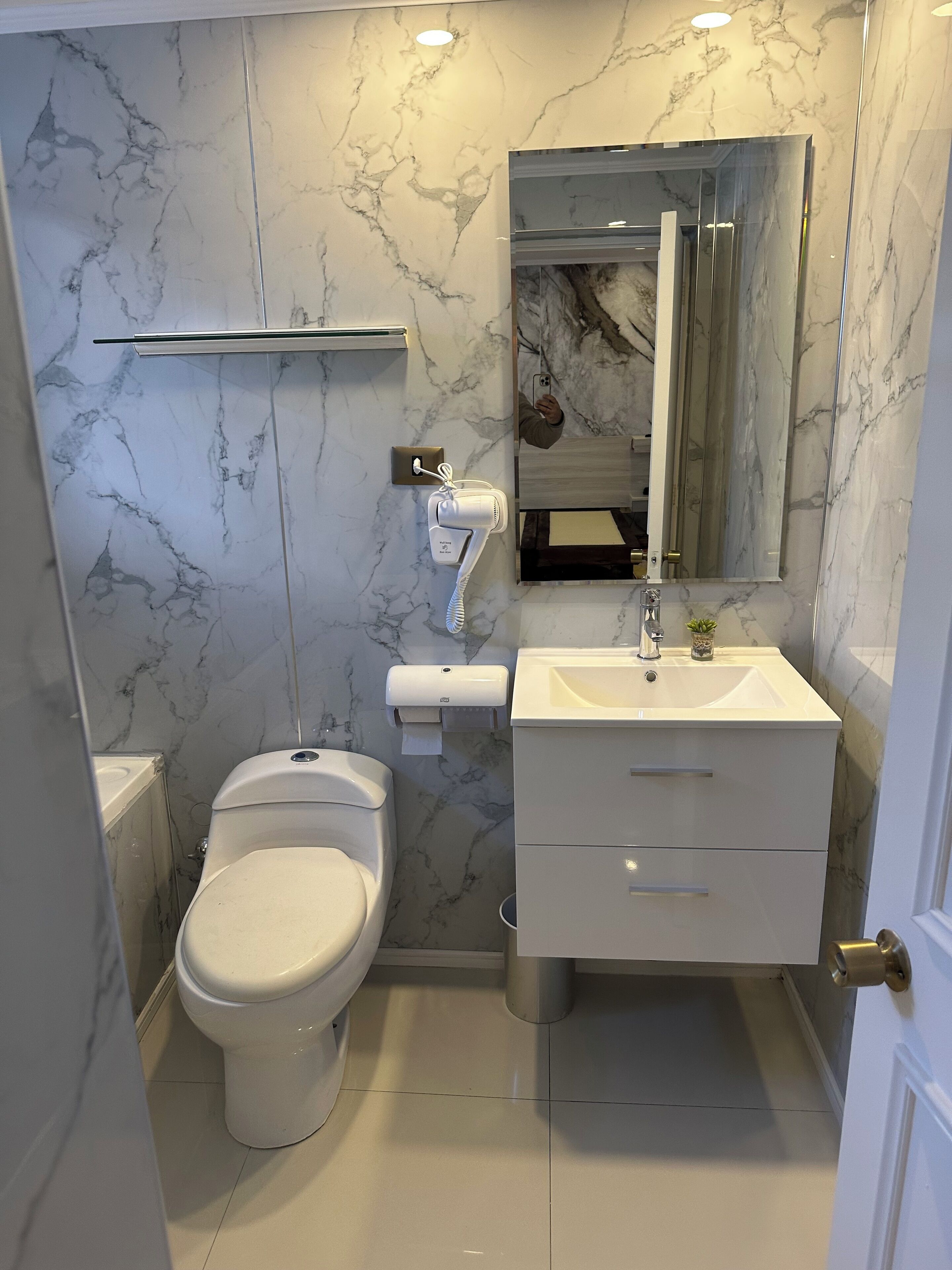 Un baño con inodoro, lavabo, espejo y secador de pelo fijado en la pared.