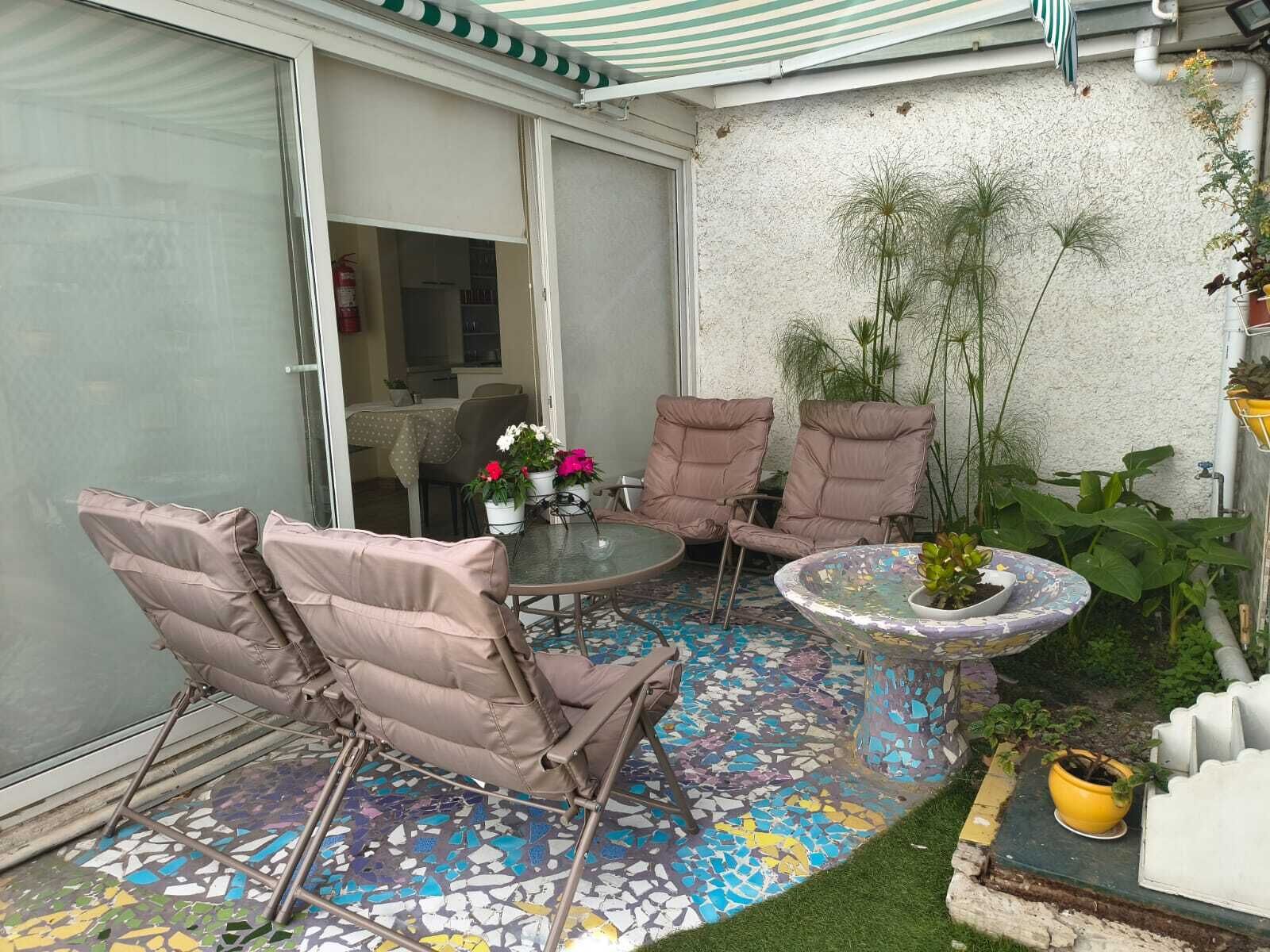 Um pátio com piso de mosaico, duas cadeiras estofadas, uma mesinha com planta em vaso e uma porta de vidro deslizante que dá para uma área interna.