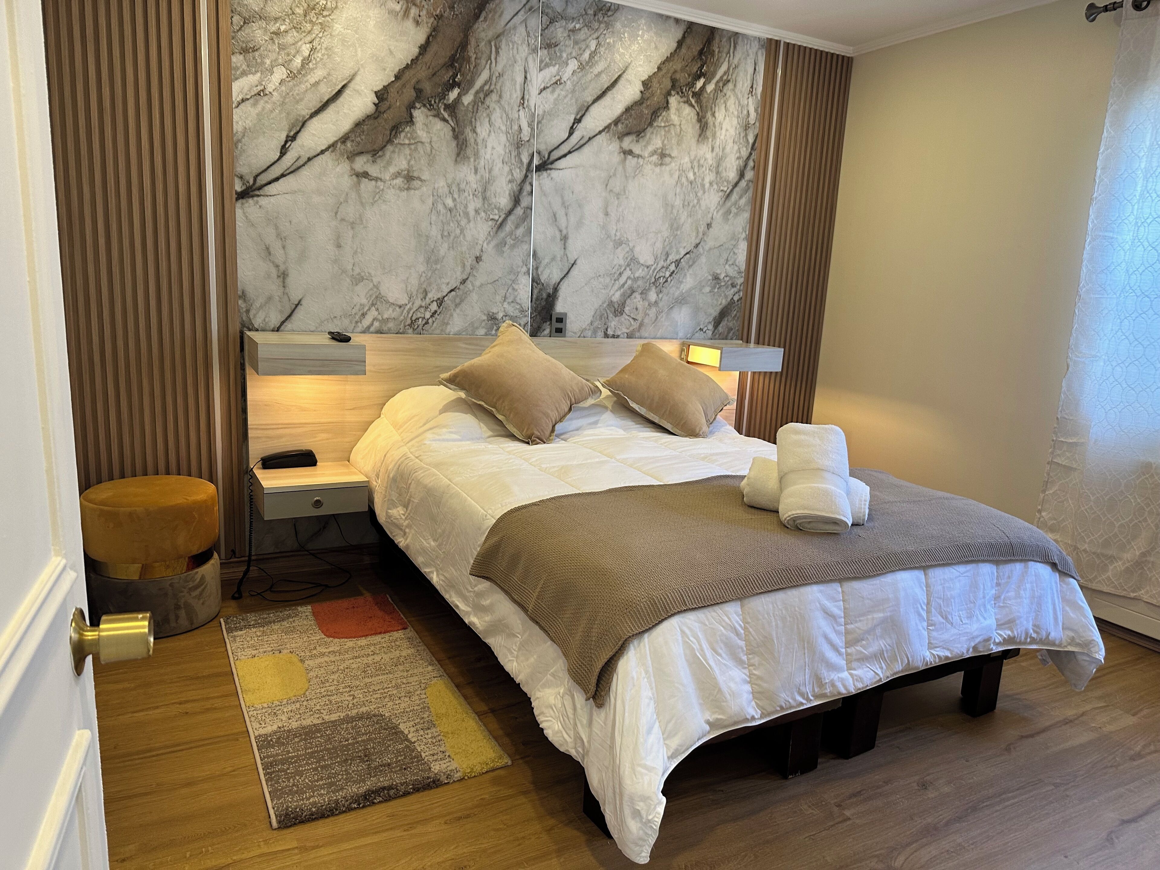 Una cama bien hecha con cabecera, una mesita de noche con lámpara, un taburete pequeño y una alfombra estampada.