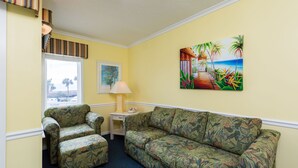 A room with a floral-patterned sofa, a matching armchair, a small table with a lamp, and a painting of a tropical scene. - 2-BR Beachfront Condo with Balcony Views! (North Myrtle Beach)
