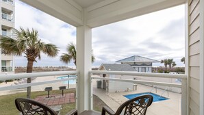 A balcony with a pool, palm trees, and a view of a building across the water. - 2-BR Beachfront Condo with Balcony Views! (North Myrtle Beach)