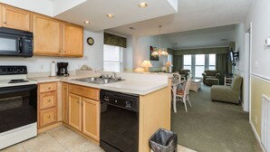 A compact kitchen with wooden cabinets, a microwave, and a dishwasher. There is a dining area with a table and chairs, and a living room with a couch and a lamp.