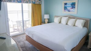 A hotel room with a large bed, a balcony with yellow curtains, a painting of a beach, and a lamp on the nightstand.