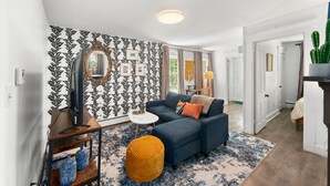 A living room with a dark blue sofa, a small round ottoman, a white side table, and a large mirror. - Charming 2-bedroom apartment near Providence (West Warwick)