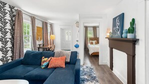 A modern living room with a blue sofa, a small table, and a fireplace. There is a bed in the background and a hallway leading to another room. - Charming 2-bedroom apartment near Providence (West Warwick)