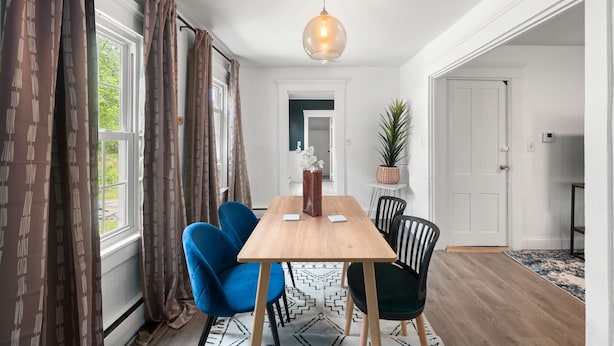 A dining area with a wooden table, blue and black chairs, a hanging light fixture, and a view of the outdoors through large windows. - Charming 2-bedroom apartment near Providence (West Warwick)