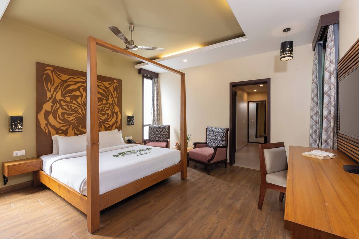 A four-poster bed with a wooden headboard, a ceiling fan, a bedside table, a chair, a wooden desk, and a wall-mounted lamp.