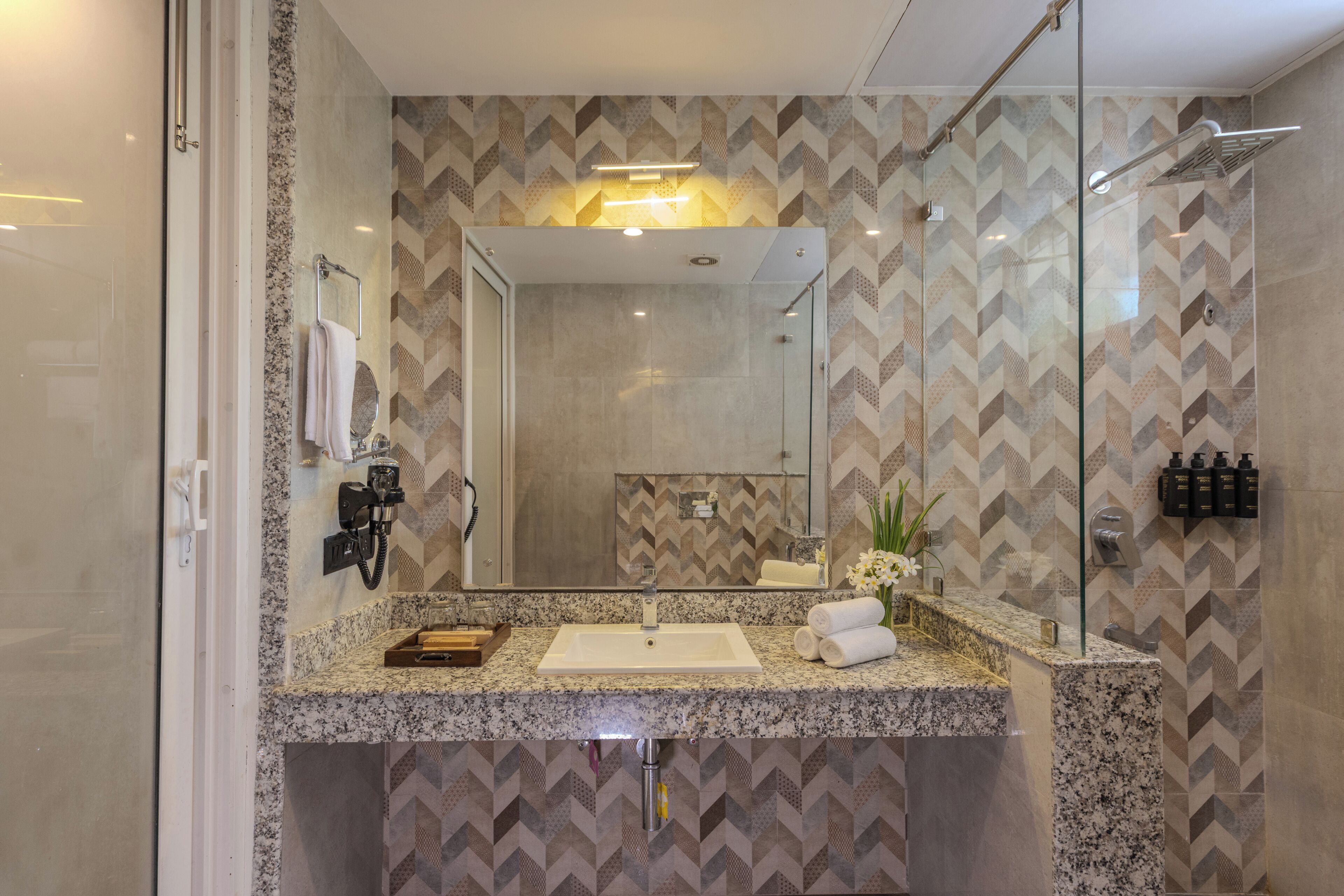 A modern bathroom with a granite countertop, a large mirror, a glass shower enclosure, and a patterned tile backsplash.