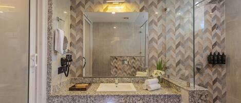 A modern bathroom with a granite countertop, a large mirror, a glass shower enclosure, and a patterned tile backsplash.