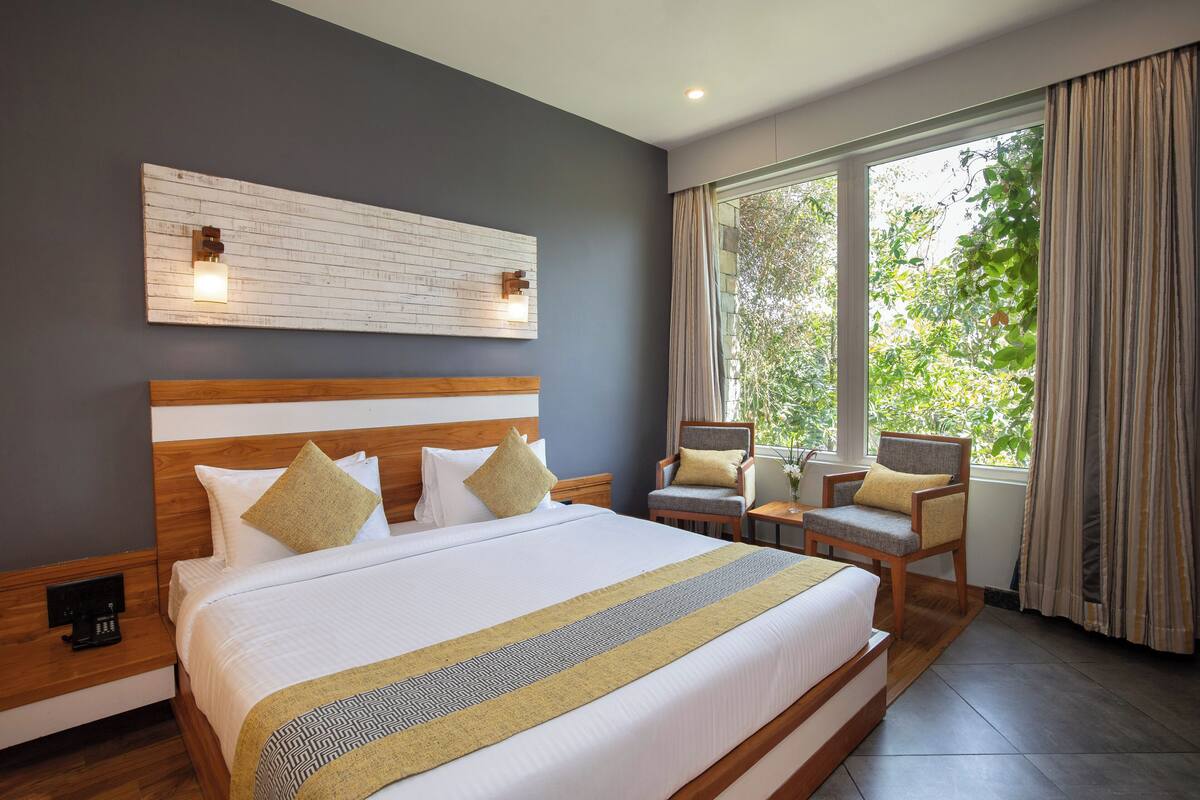 A hotel room with a large bed, a wooden headboard, a wall-mounted light fixture, a window with curtains, and a chair with a cushion.