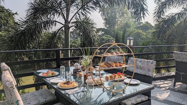 A glass-top dining table with a tiered dessert stand, surrounded by wicker chairs and a view of lush greenery. - The Golden Tusk By Ivory Destinations (Ramnagar)