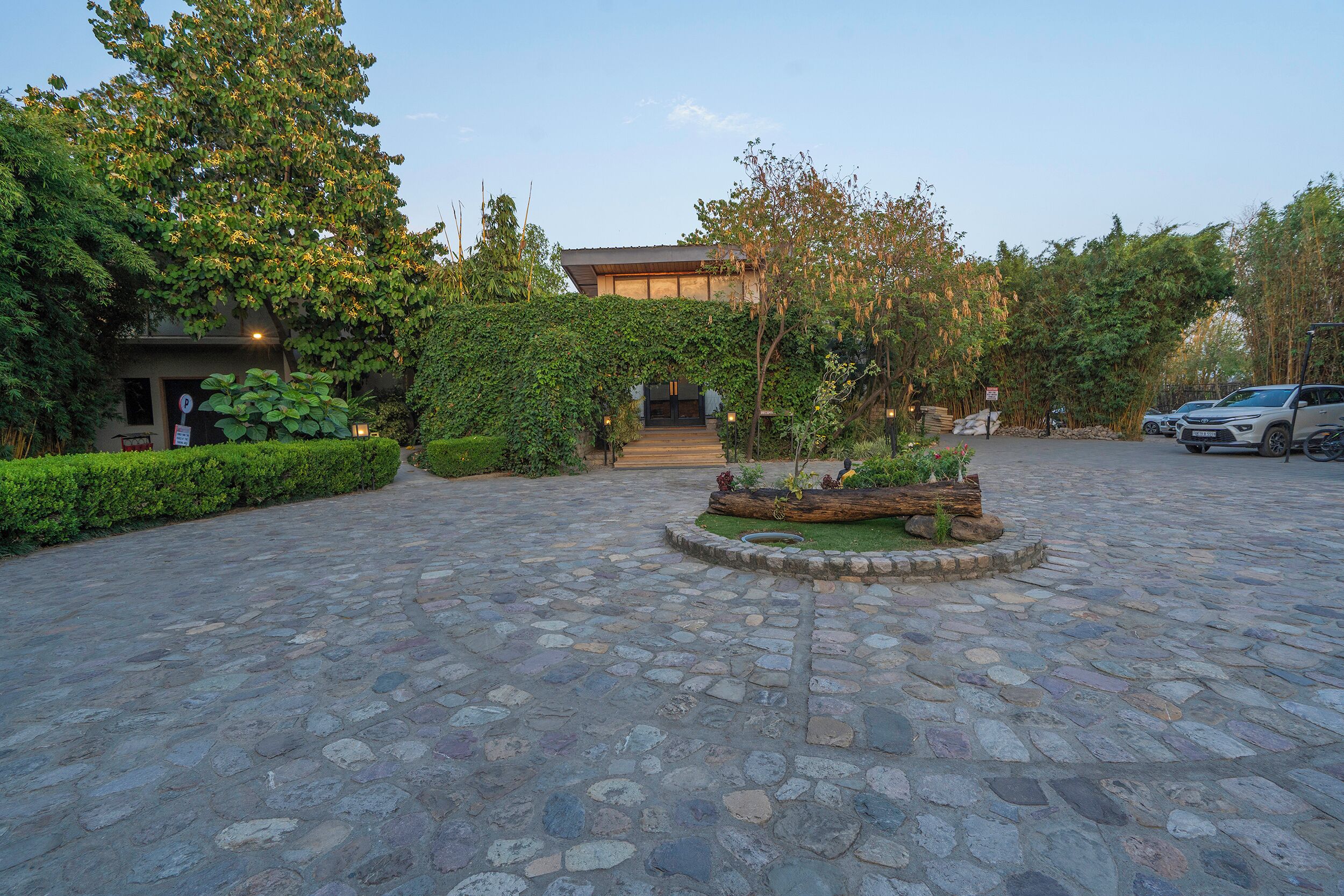 A courtyard with a stone-paved ground, a central garden with a wooden log and plants, and a building with a dark roof in the background.