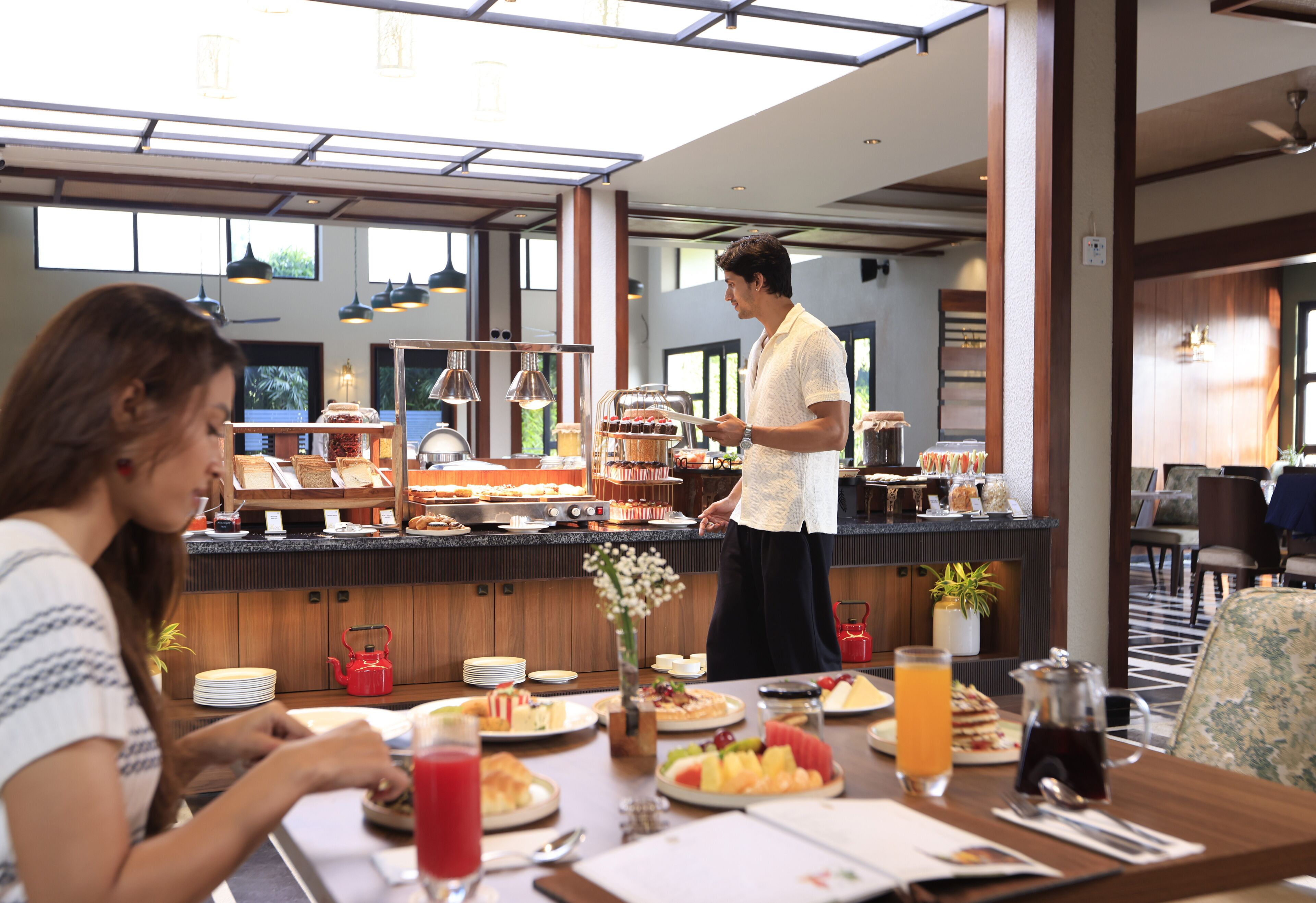 Ein reichhaltiges Frühstücksbuffet mit zahlreichen Speisen, ein Kellner bedient Gäste, und ein modernes Interieur mit großen Fenstern und Pendelleuchten.