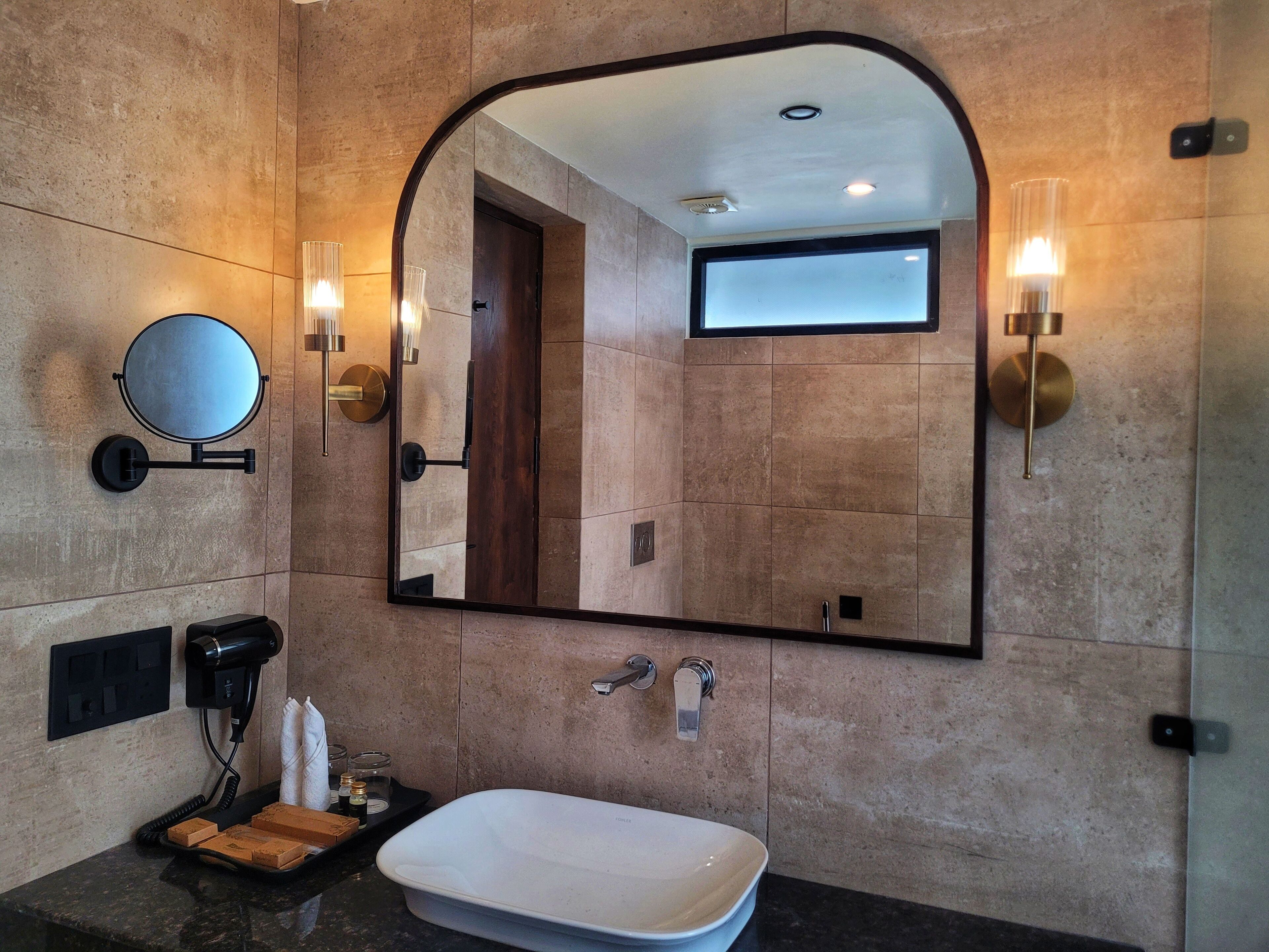Un baño moderno con un espejo grande, un espejo redondo, un lavamanos rectangular e iluminación en la pared.