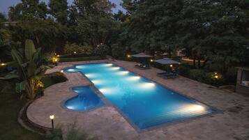 Una piscina rectangular con un jacuzzi pequeño, rodeada por un solárium empedrado y abundante vegetación.