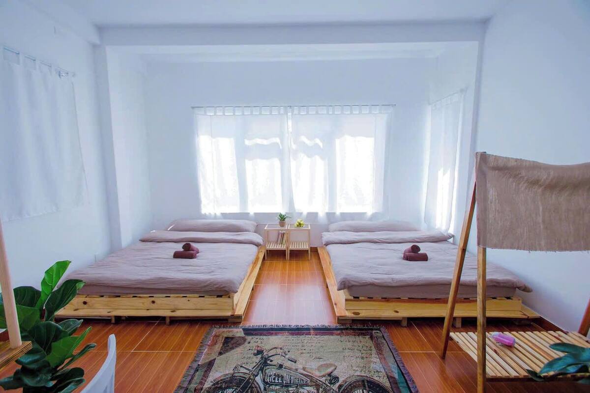 A room with two beds, a small table, and a window with sheer curtains.