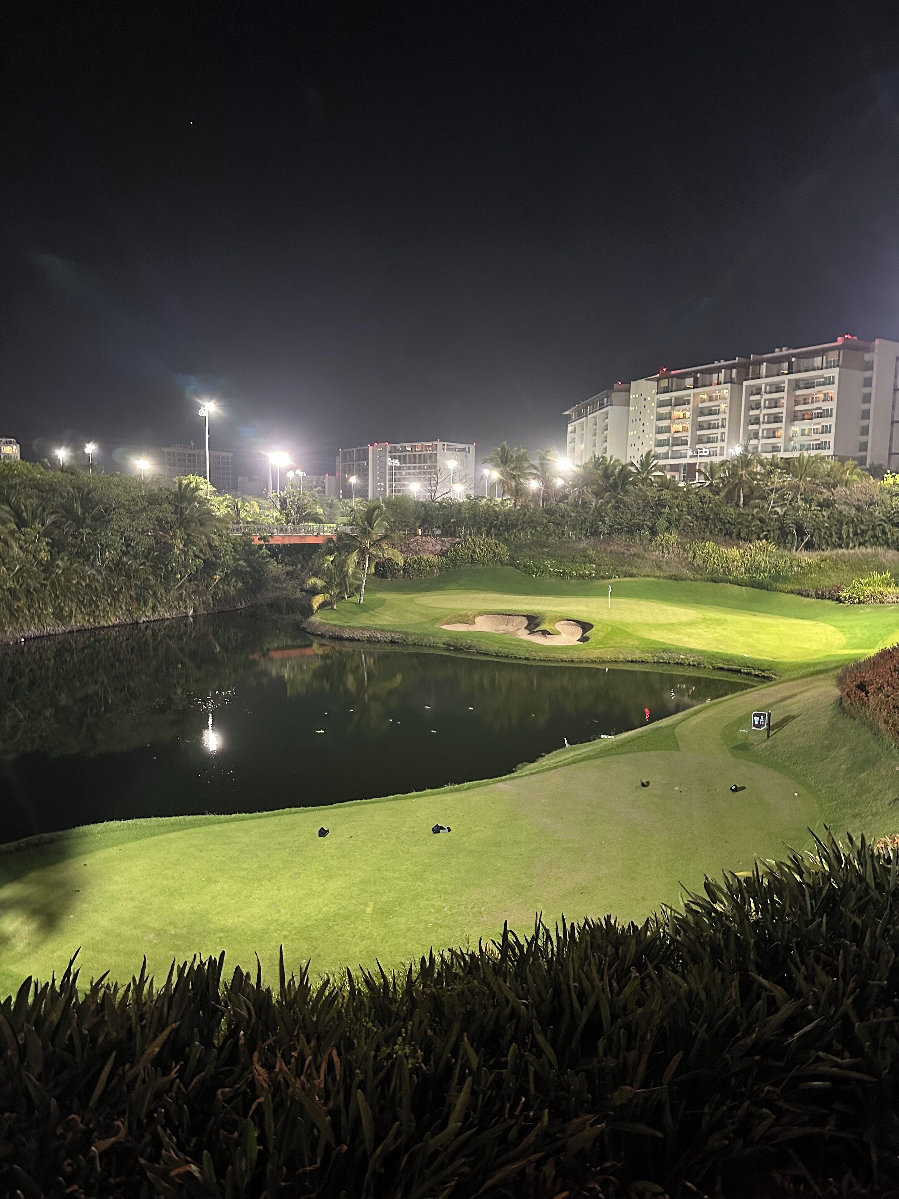 A nighttime view of a golf course with a pond, surrounded by well-lit buildings and lush greenery.