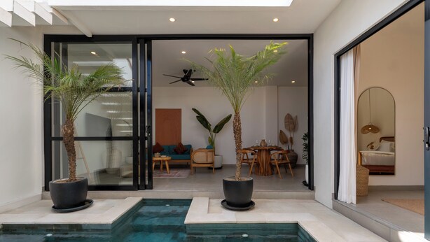 A modern indoor pool area with two palm trees, a ceiling fan, and a view into a living room with a sofa and dining table. - Enaya Villa by Betterplace (Canggu)