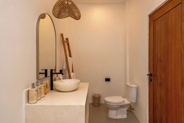A bathroom with a large mirror, a sink with a bowl, a toilet, and a wooden door. - Enaya Villa by Betterplace (Canggu)
