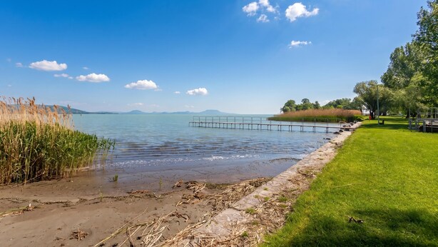 Vista de un lago con una zona verde, un muelle y montañas a lo lejos.