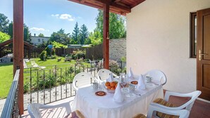 Un patio techado al aire libre con una mesa lista para comer, rodeado de sillas blancas, y una vista de un jardín y edificios al fondo.