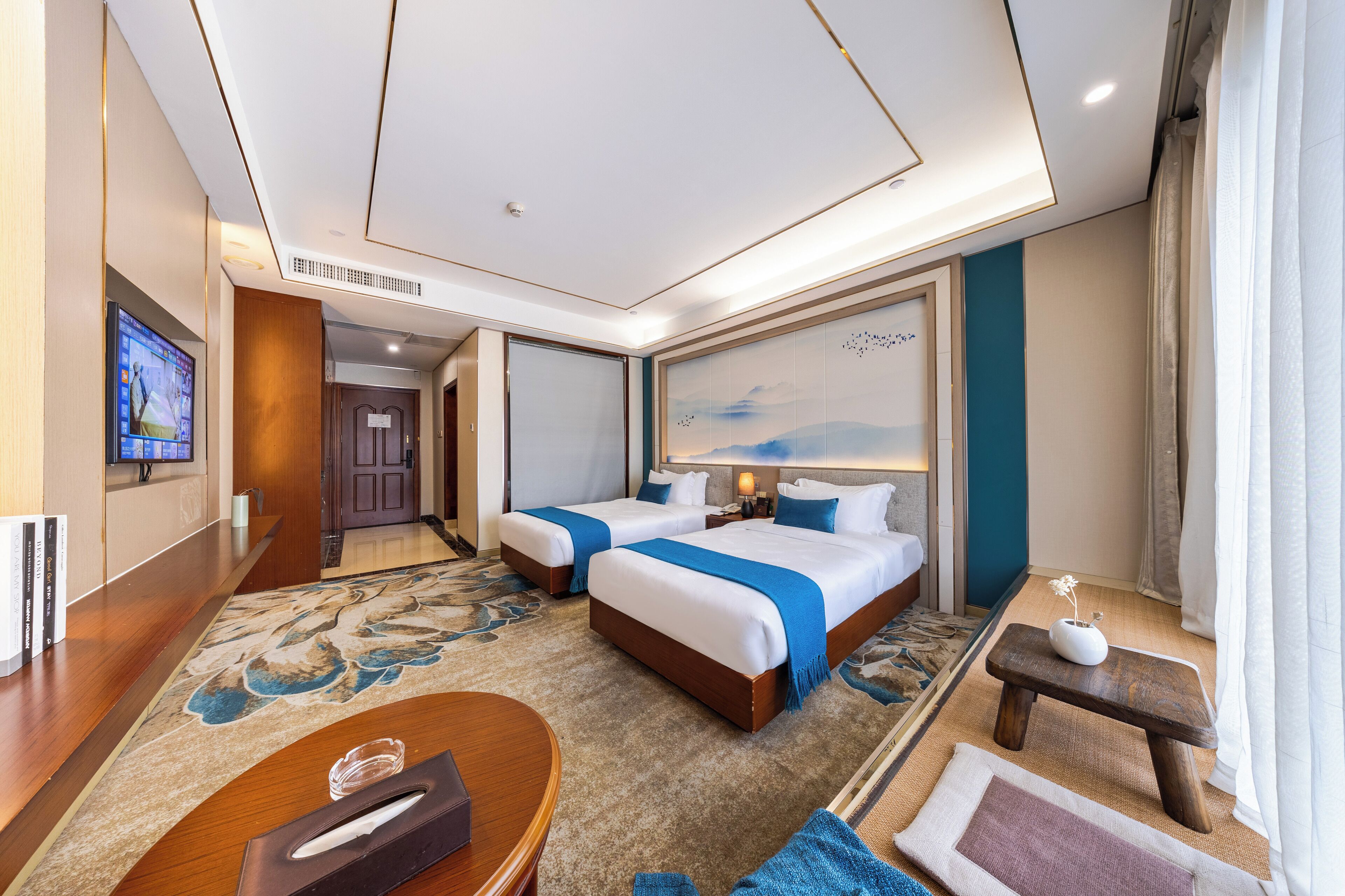 A hotel room with two beds, a TV, a desk, and a small table with a vase.