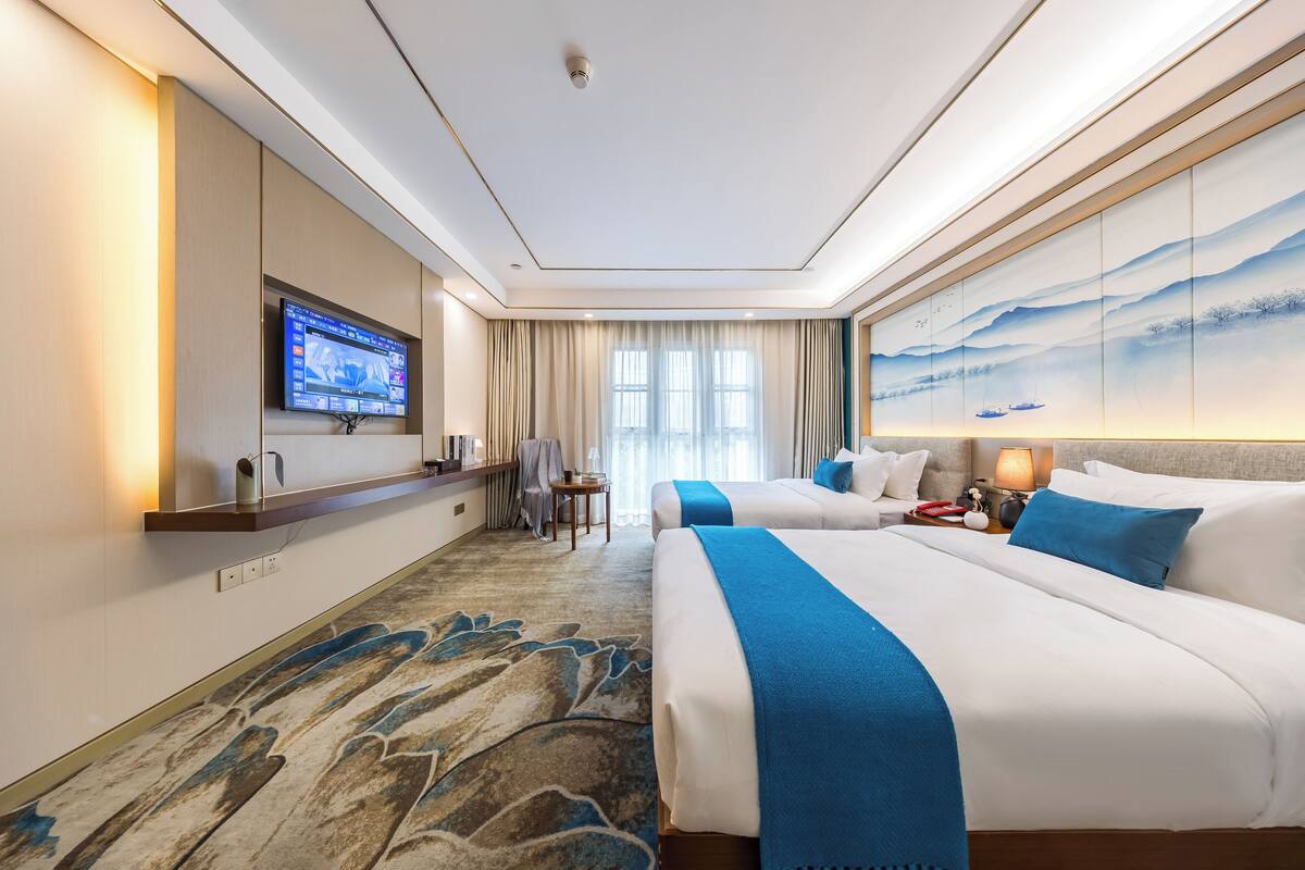 Habitación de hotel con dos camas, un televisor de pantalla plana montado en la pared, un ventanal con cortinas y un cuadro decorativo en la pared.